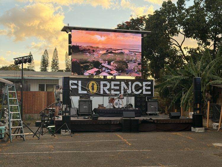 Outdoor concert stage with 20-ft LED wall showing a Haleʻiwa sunset aerial, Florence banner